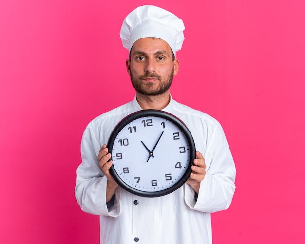 Serious young caucasian male cook in chef uniform and cap holding clock 