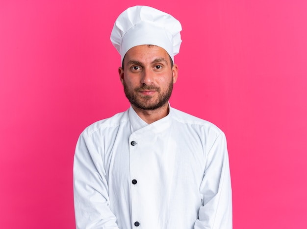 Grave giovane maschio caucasico cuoco in uniforme da chef e berretto guardando la telecamera isolata sulla parete rosa