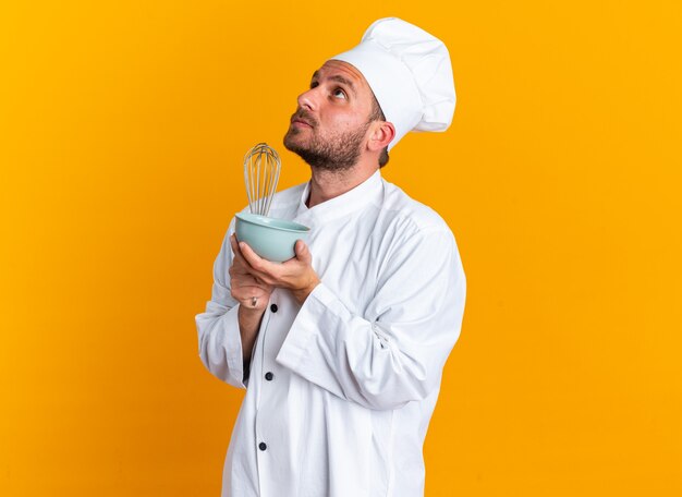 Serious young caucasian male cook in chef uniform and cap standing in profile view holding whisk and bowl looking up isolated on orange wall with copy space