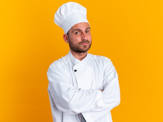 Serious young caucasian male cook in chef uniform and cap standing with closed posture looking at camera isolated on orange wall