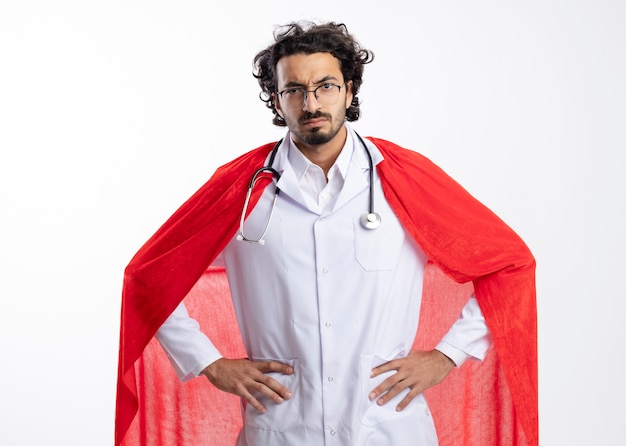 Free photo serious young caucasian superhero man in optical glasses wearing doctor uniform with red cloak and with stethoscope around neck stands putting hands on waist isolated on white wall