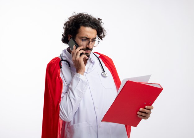 Serious young caucasian superhero man in optical glasses wearing doctor uniform with red cloak and with stethoscope around neck talking on phone looking at file folder with copy space