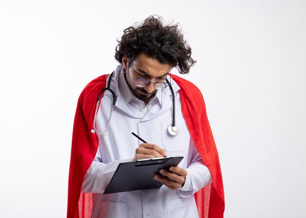 Serious young caucasian superhero man in optical glasses wearing doctor uniform with red cloak and with stethoscope around neck writes with pencil in clipboard on white wall with copy space