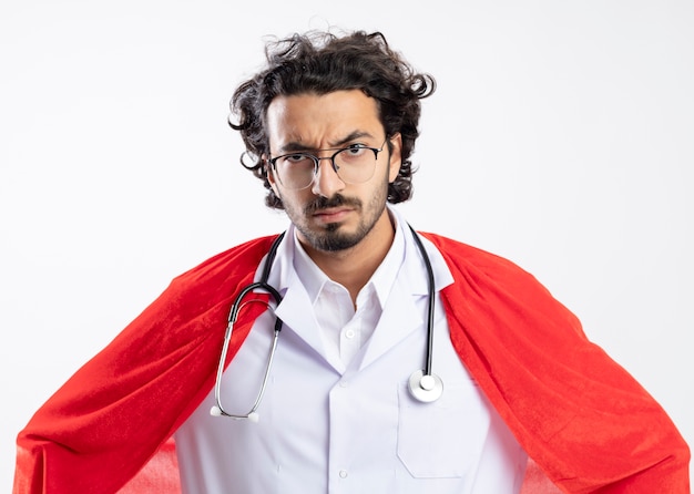 Free photo serious young caucasian superhero man in optical glasses wearing doctor uniform with red cloak and with stethoscope around neck