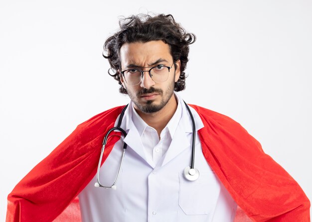 Serious young caucasian superhero man in optical glasses wearing doctor uniform with red cloak and with stethoscope around neck 