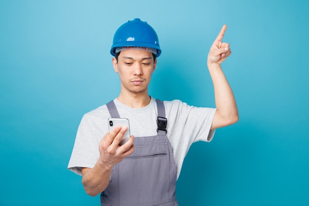 Serious young construction worker wearing safety helmet and uniform holding and looking at mobile phone pointing up 