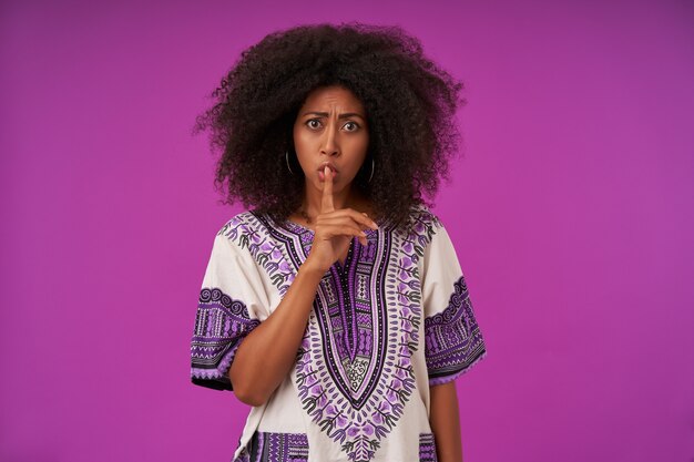 Serious young dark skinned lady with casual hairstyle posing on purple, frowning and raising index finger in hush gesture, wearing white patterned shirt