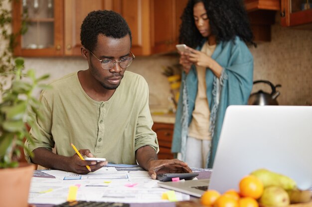 Serious young dark-skinned male in spectacles using cell phone and calculator while calculating family expenses