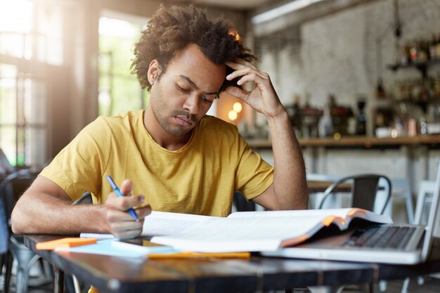 Серьезный молодой темнокожий мужчина с темными волосами и щетиной в желтой футболке, сосредоточенно глядя в блокнот, готовясь к экзамену или урокам, сидя в кафетерии, усердно работает