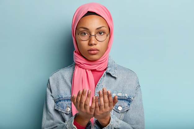Serious young dark skinned woman looks at camera, prays indoor, keeps two hands in praying gesture
