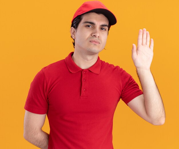 Serious young delivery man in blue uniform and cap keeping hand behind back looking at front showing empty hand isolated on orange wall