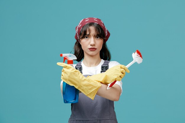 青い背景で隔離のカメラを見て交差したブラシとクレンザーを保持している制服のバンダナとゴム手袋を身に着けている深刻な若い女性クリーナー