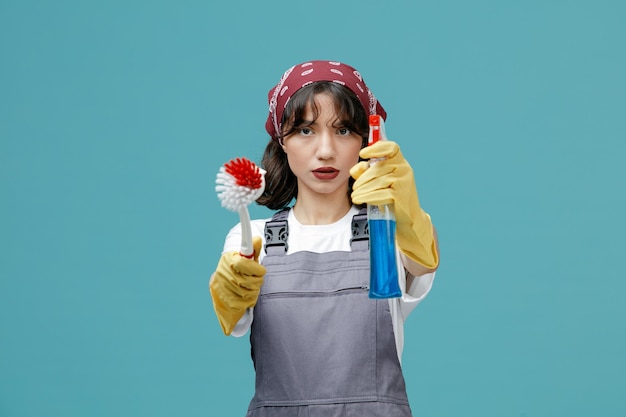 Serious young female cleaner wearing uniform bandana and rubber gloves looking at camera stretching brush and cleanser out towards camera isolated on blue background