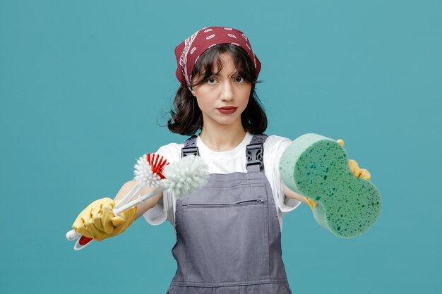 Free photo serious young female cleaner wearing uniform bandana and rubber gloves looking at camera stretching sponge and toilet brushes out towards camera isolated on blue background