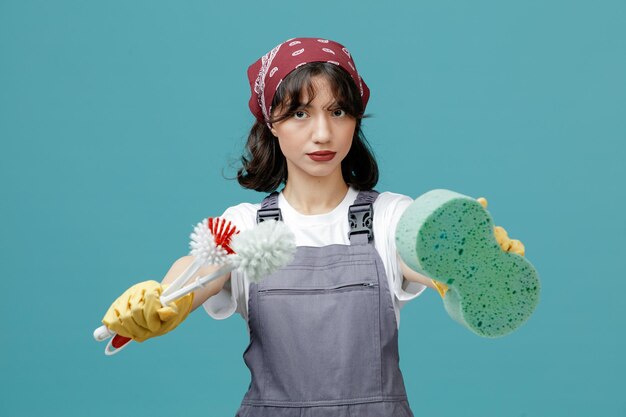 serious young female cleaner wearing uniform bandana and rubber gloves looking at camera stretching sponge and toilet brushes out towards camera isolated on blue background
