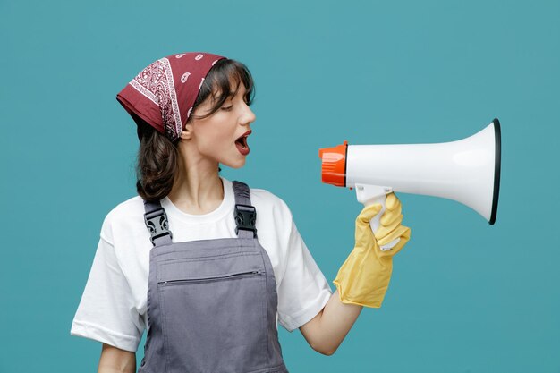 Serious young female cleaner wearing uniform bandana and rubber gloves looking at side talking into speaker isolated on blue background