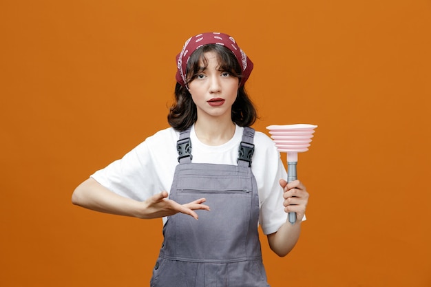 Serious young female cleaner wearing uniform and bandana showing plunger pointing at it with hands looking at camera isolated on orange background