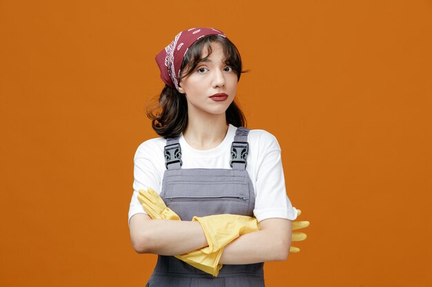 Serious young female cleaner wearing uniform rubber gloves and bandana keeping arms crossed looking at camera isolated on orange background