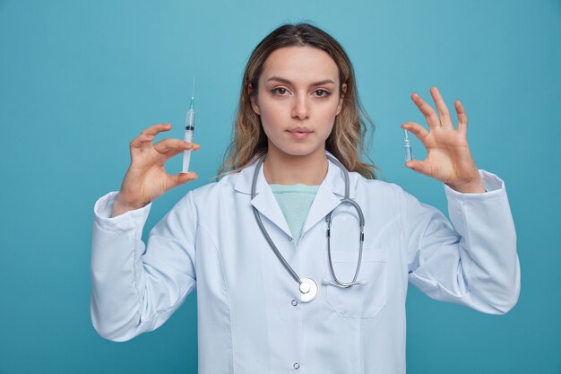 Serious young female doctor wearing medical robe and stethoscope around neck holding syringe and ampoule 