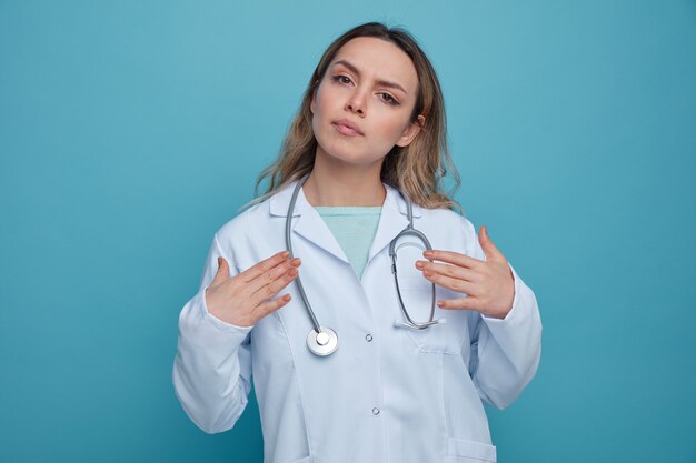 Серьезная молодая женщина-врач в медицинском халате и стетоскопе на шее, указывая на себя руками