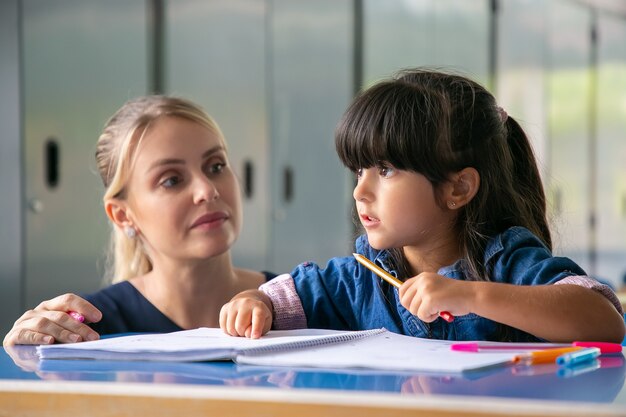 Серьезная молодая учительница помогает девочке начальной школы выполнять свою задачу