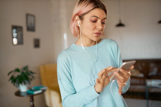 Serious young female with facial piercing working distantly using cell phone, listening to music in earphones