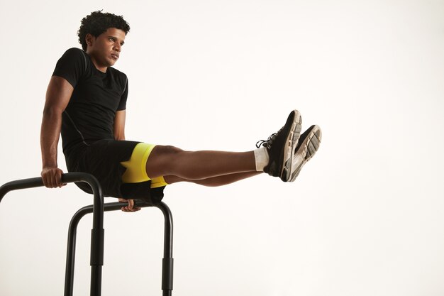 Serious young fit African American man in black sportswear doing L-sits on short bars at home against a white wall