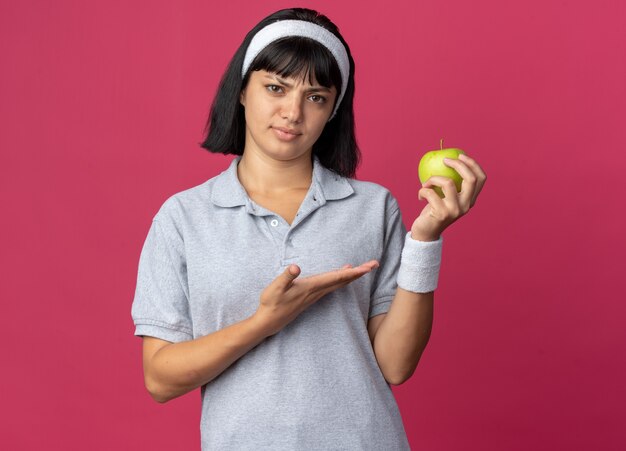 Serious young fitness girl wearing headband holding green apple looking at camera presenting it with arm 