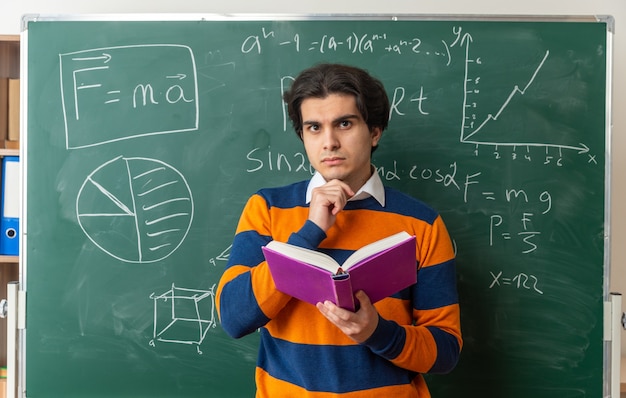 真面目な若い幾何学教師が教室の黒板の前に立って本を持って正面を見てあごに手を置いている