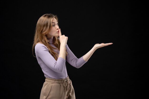 Serious young girl pointing something on the left side and making silence gesture on dark