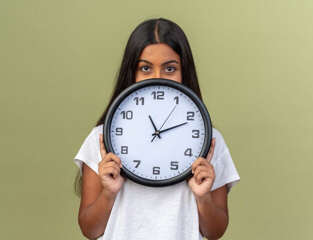 Serious young girl in white t-shirt holding wall clock hiding her face behind it 