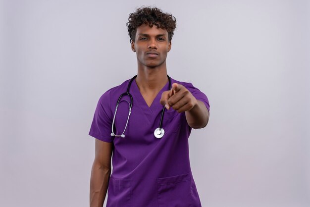 Серьезный молодой красивый темнокожий мужчина-врач с вьющимися волосами в фиолетовой форме со стетоскопом, указывающим указательным пальцем на камеру на белом фоне