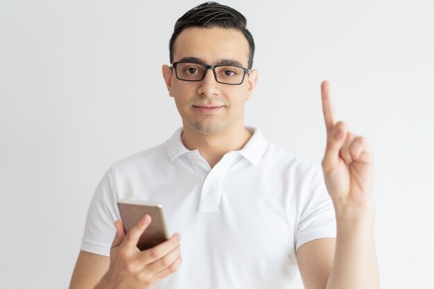 Serious young guy pointing upwards and holding smartphone