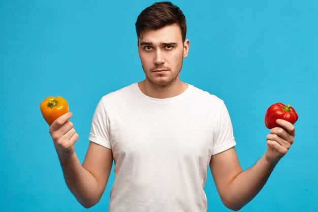 Серьезный молодой парень с щетиной, держащий апельсин и красный сладкий перец с нерешительным выражением лица, имеющий сомнения и подозрения, не хочет есть овощи с пестицидами