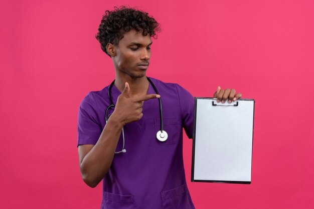 A serious young handsome dark-skinned doctor with curly hair wearing violet uniform with stethoscope pointing with index finger at clipboard with blank sheet of paper 