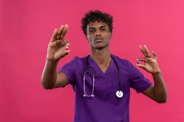 A serious young handsome dark-skinned doctor with curly hair wearing violet uniform with stethoscope showing pills 