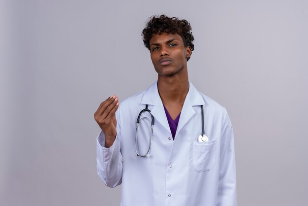 A serious young handsome dark-skinned male doctor with curly hair wearing white coat with stethoscope raising hand 