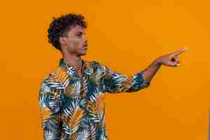Free photo serious young handsome dark-skinned man with curly hair in leaves printed shirt pointing with index finger on an orange background