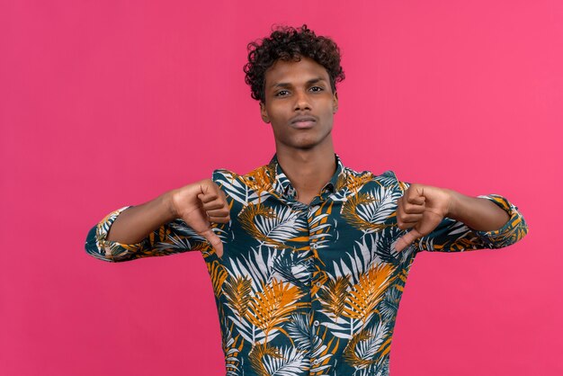 A serious young handsome dark-skinned man with curly hair in leaves printed shirt with a dissentingseriousstern expression with thumbs down 