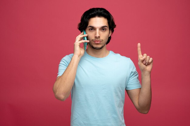 赤い背景で隔離の電話で話しているカメラを上向きに見ている深刻な若いハンサムな男