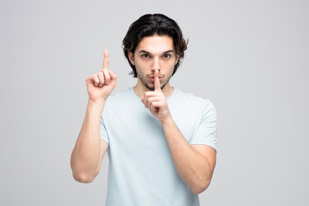 Serious young handsome man looking at camera showing silence gesture pointing up isolated on white background