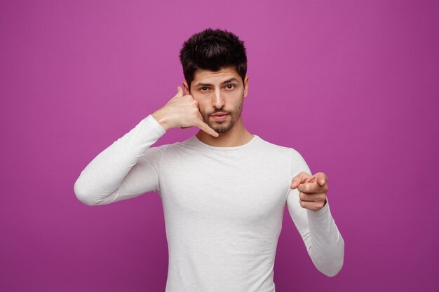 Serious young handsome man looking and pointing at camera doing phone gesture on purple background