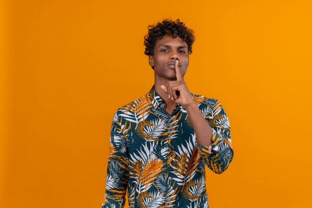 Serious young handsome man showing shh gesture with index finger near mouth on an orange background
