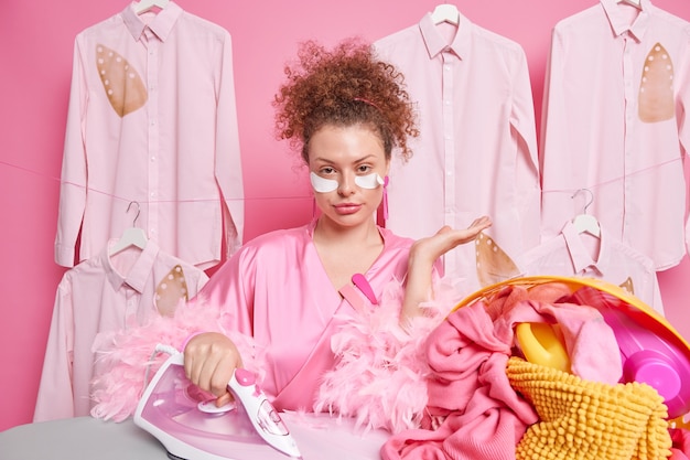 Free photo serious young housewife wears domestic gown busy ironing laundry undergoes beauty procedures at home poses against ironed shirts on hangers busy with household chores. domestic life concept.