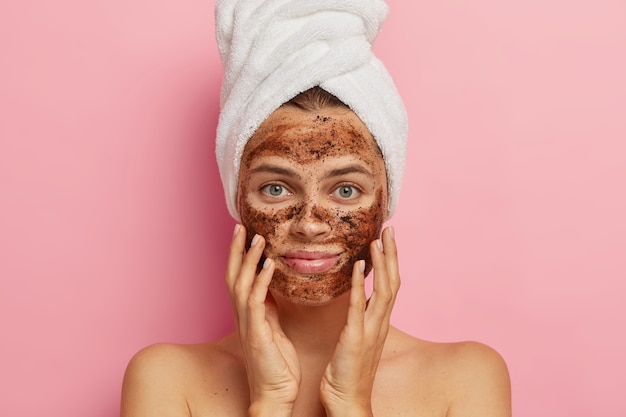 Free photo serious young lady applies coffee scrub on face, does peeling of skin, removes pores, touches cheeks with hands, has naked body