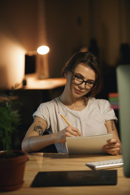 Serious young lady designer sitting indoors at night writing notes