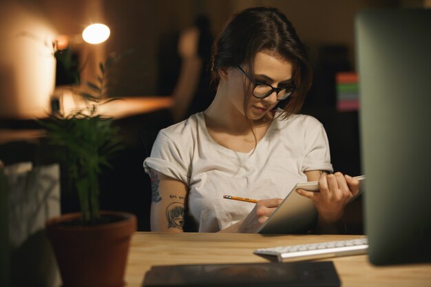 Serious young lady designer sitting indoors at night writing notes.