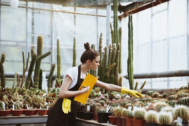 Serious young lady standing in greenhouse holding clipboard