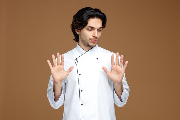 серьезный молодой шеф-повар в униформе смотрит вниз, не показывая жеста на коричневом фоне