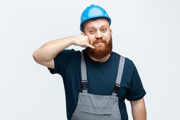 Serious young male construction worker wearing safety helmet and uniform looking at camera showing call me gesture isolated on white background